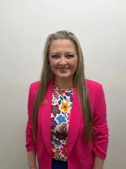 A woman with brown hair and blue eyes smiling, standing in front of a red brick wall. She is wearing a blue button-down shirt, a white scarf, and colorful yellow and blue earrings.