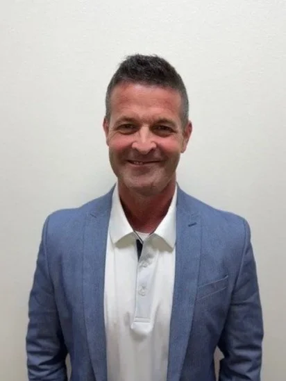 A man in a light blue dress shirt and pink patterned tie standing against a red brick wall, smiling at the camera.