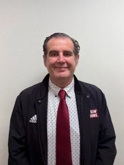 A man with dark, curly hair and a mustache, wearing a navy blue shirt and a patterned tie, smiling in front of a red brick wall.