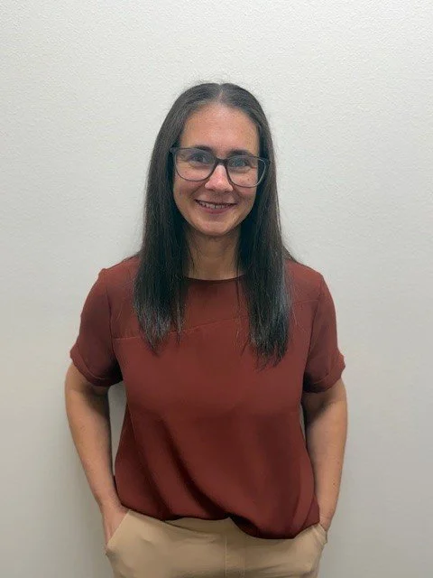 A woman with glasses and wavy brown hair standing in front of a brick wall, wearing a navy blue shirt and a patterned scarf.