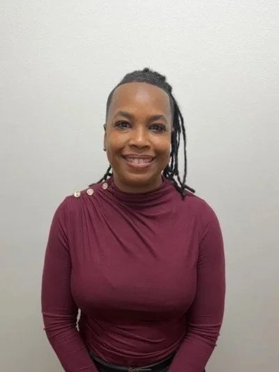 A woman with short dreadlocks, wearing a light blue button-up shirt, white pants, a brown belt, and a gold watch, standing outside against a red brick wall and smiling.