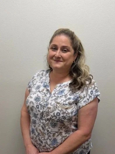 A woman standing with her hands behind her back in front of a brick wall, wearing a light purple blouse and a floral scarf.