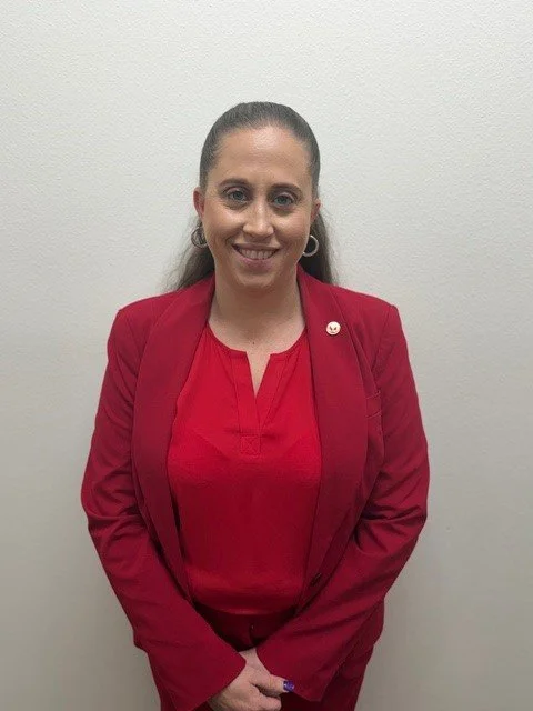 A woman with long brown hair in a ponytail, wearing a royal blue blazer and silver hoop earrings, stands next to a brick wall outside, smiling at the camera.