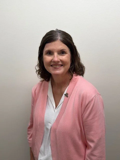 A smiling woman with dark hair in a ponytail, wearing a gray fleece jacket, beige shirt, and blue bandana, standing in front of a red brick wall.
