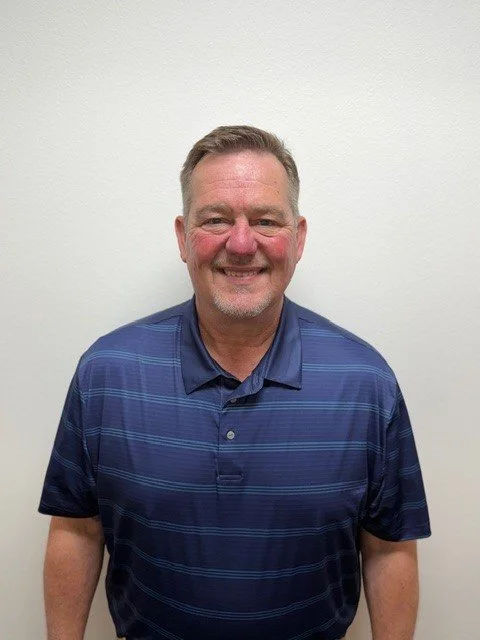 A man with short brown hair, wearing a light blue collared shirt under a dark blue Columbia fleece jacket, standing in front of a brick wall with various shades of red and brown bricks.