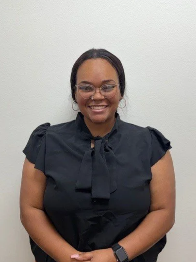 A woman with curly blonde hair, wearing glasses, smiling, stands next to a brick wall in an outdoor setting.
