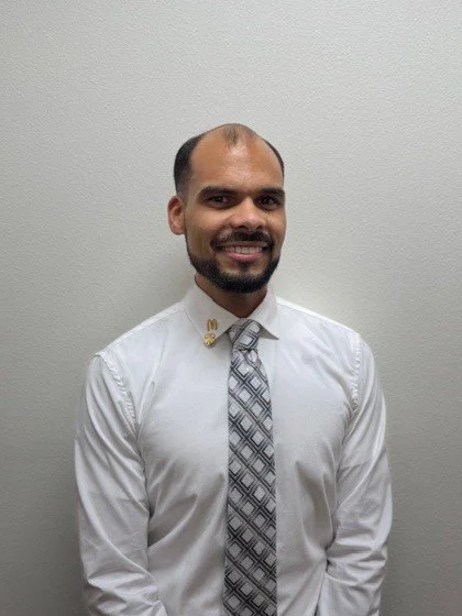 A man with a beard and mustache wearing a light blue dress shirt and a patterned tie, standing in front of a brick wall.