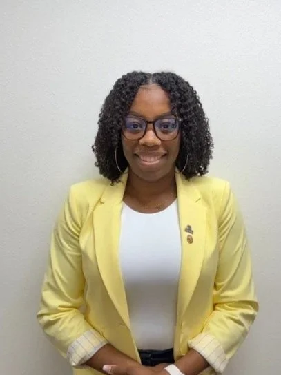 A woman smiling in front of a red brick wall, wearing a yellow blazer, a white shirt, a patterned blue and green scarf, and earrings.