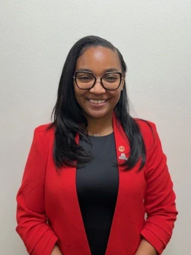 A young woman with glasses and curly black hair, wearing a white uniform shirt with a yellow and gray scarf, standing against a red brick wall and smiling at the camera.
