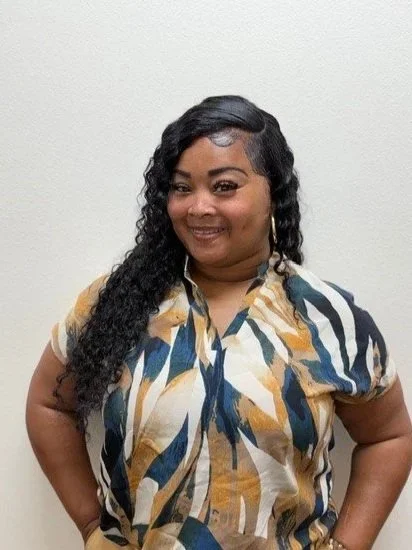 A woman wearing a yellow shirt and a patterned scarf around her neck, standing in front of a brick wall, smiling.