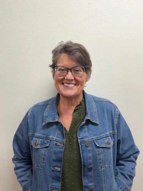 A woman with glasses, smiling, wearing a blue shirt and a patterned scarf, standing against a brick wall.