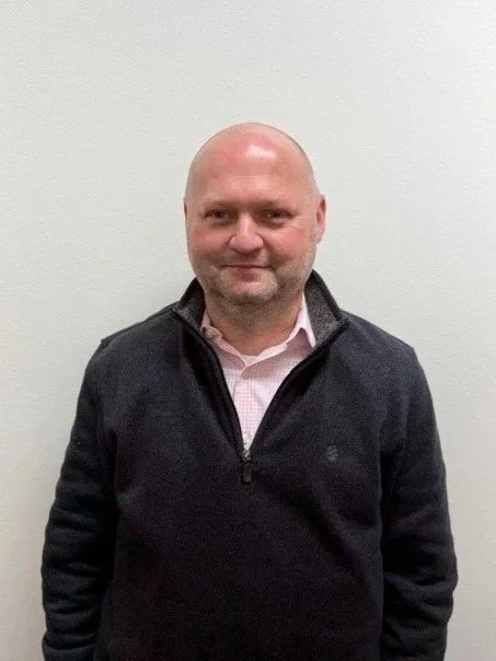 A man with a bald head and light skin standing outdoors against a brick wall, wearing a blue and white checkered button-down shirt.