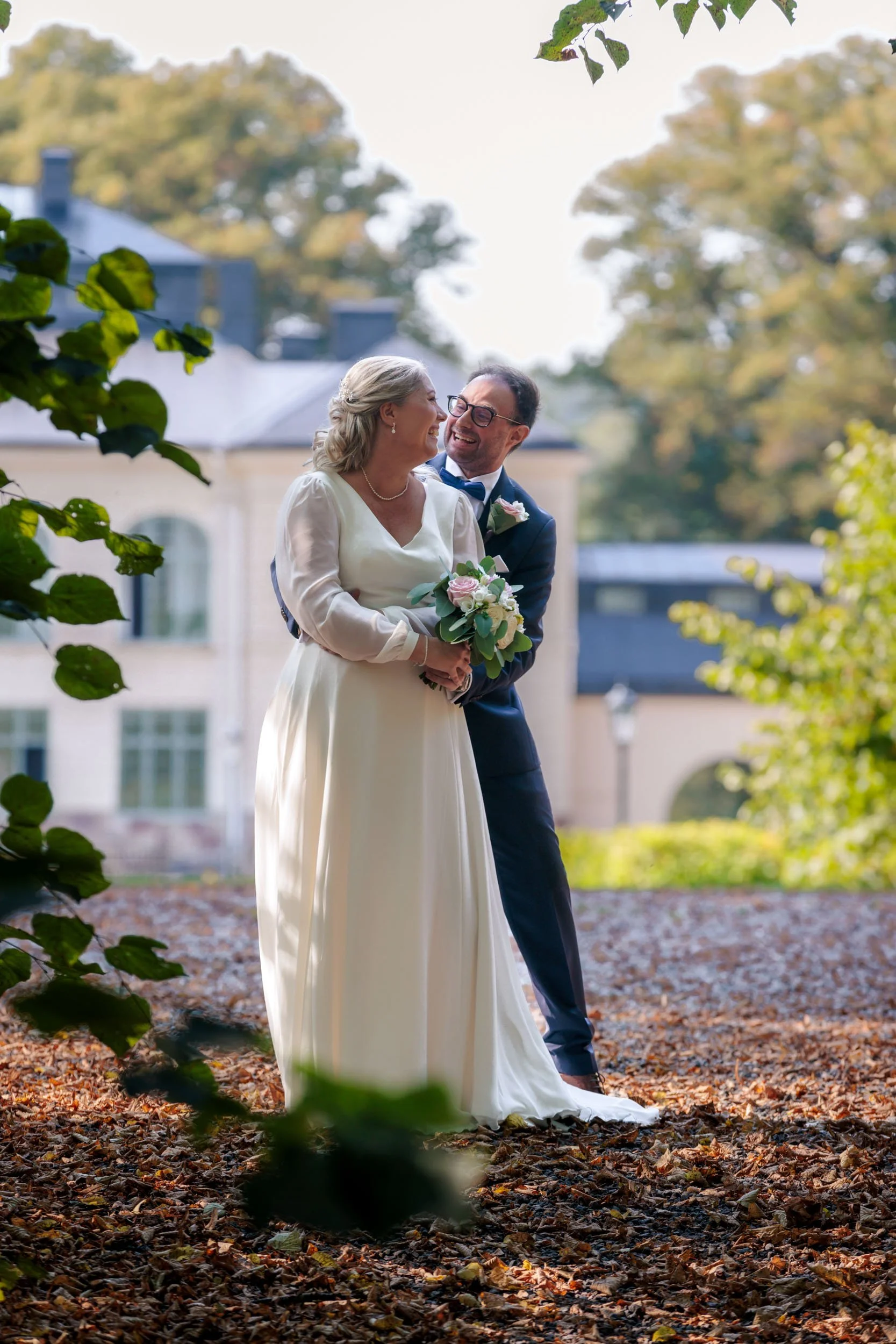 En brud och en groom som ler mot varandra utomhus, omgivna av höstlöv och träd, med ett gammalt hus i bakgrunden.