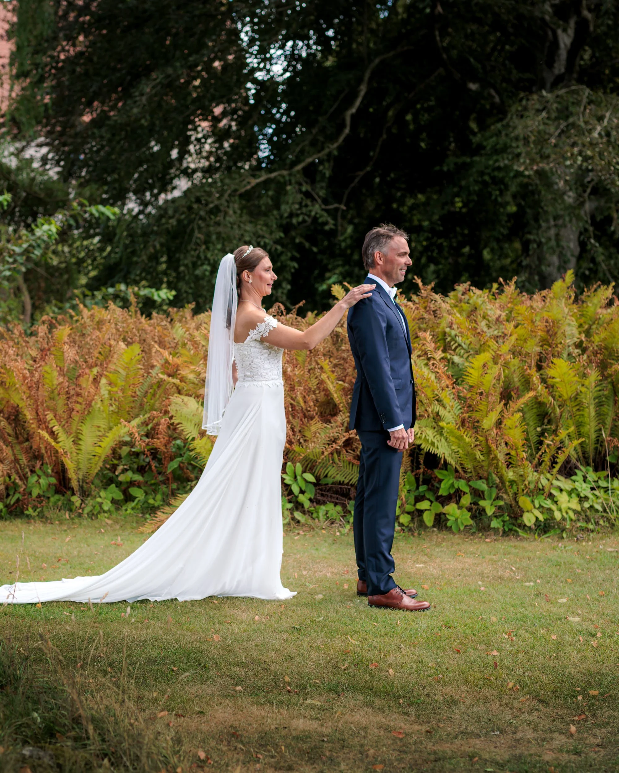 En brud i vit bröllopsklänning och en man i mörk blå kostym står i en trädgård med höstlöv på marken.