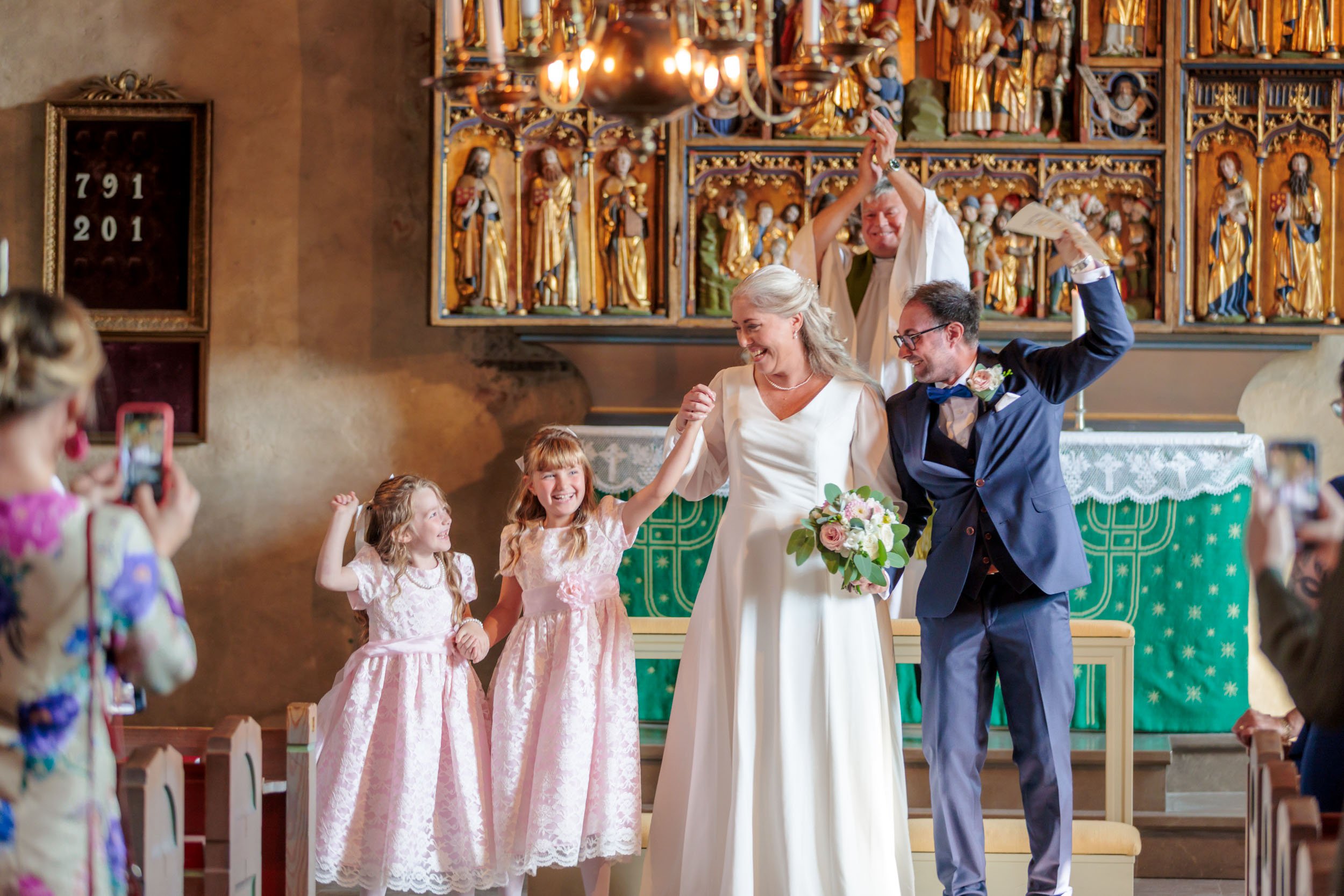 Vigsel i kyrka med ett par som gifter sig, omgivna av barn och gäster. Alla ler och ser glada ut, några tar bilder. Kyrkans altare syns i bakgrunden med religiöst dekorerad vägg.