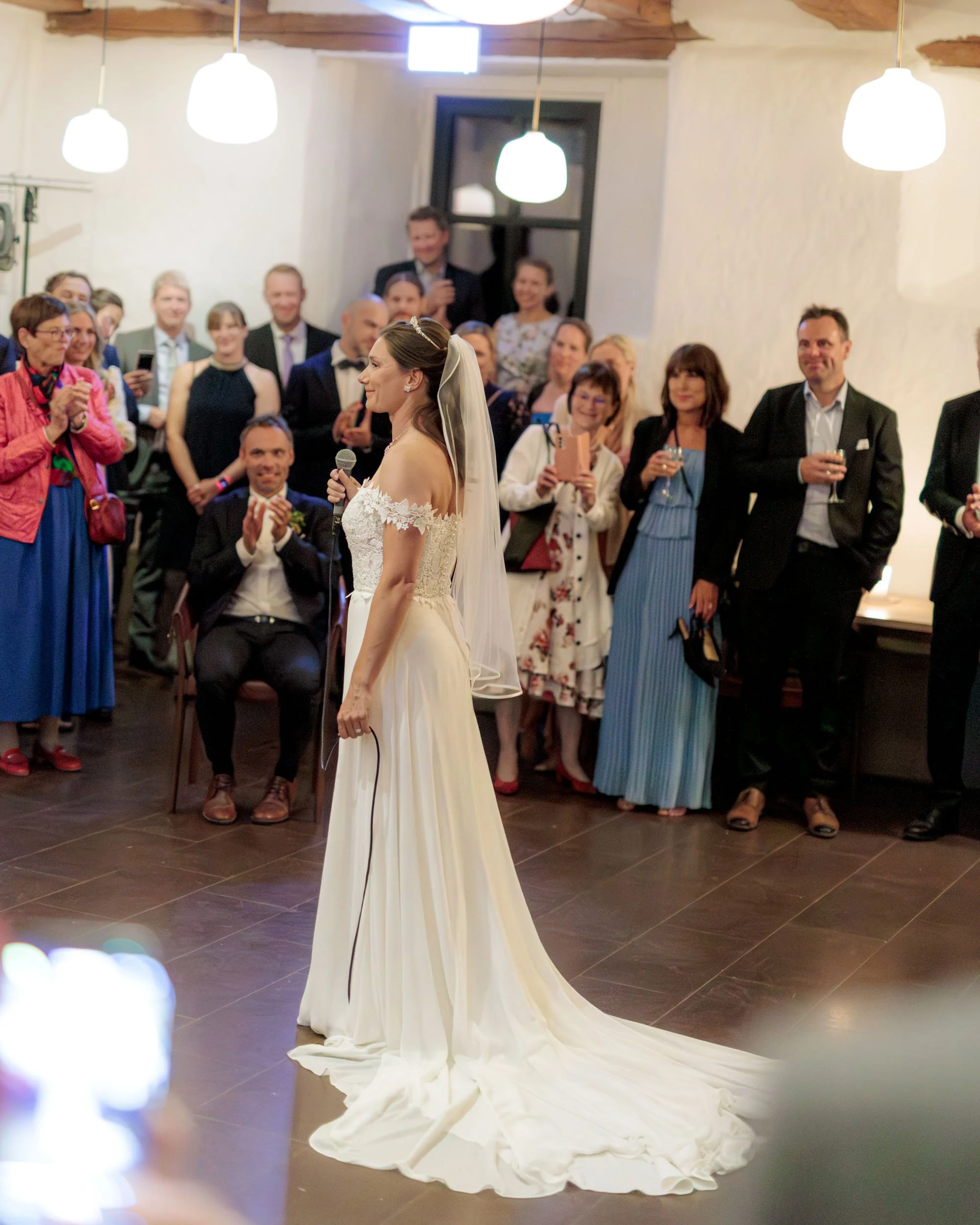 Brudklädd kvinna håller mikrofon och talar i ett bröllop, omgiven av gäster som ser på och fotograferar.