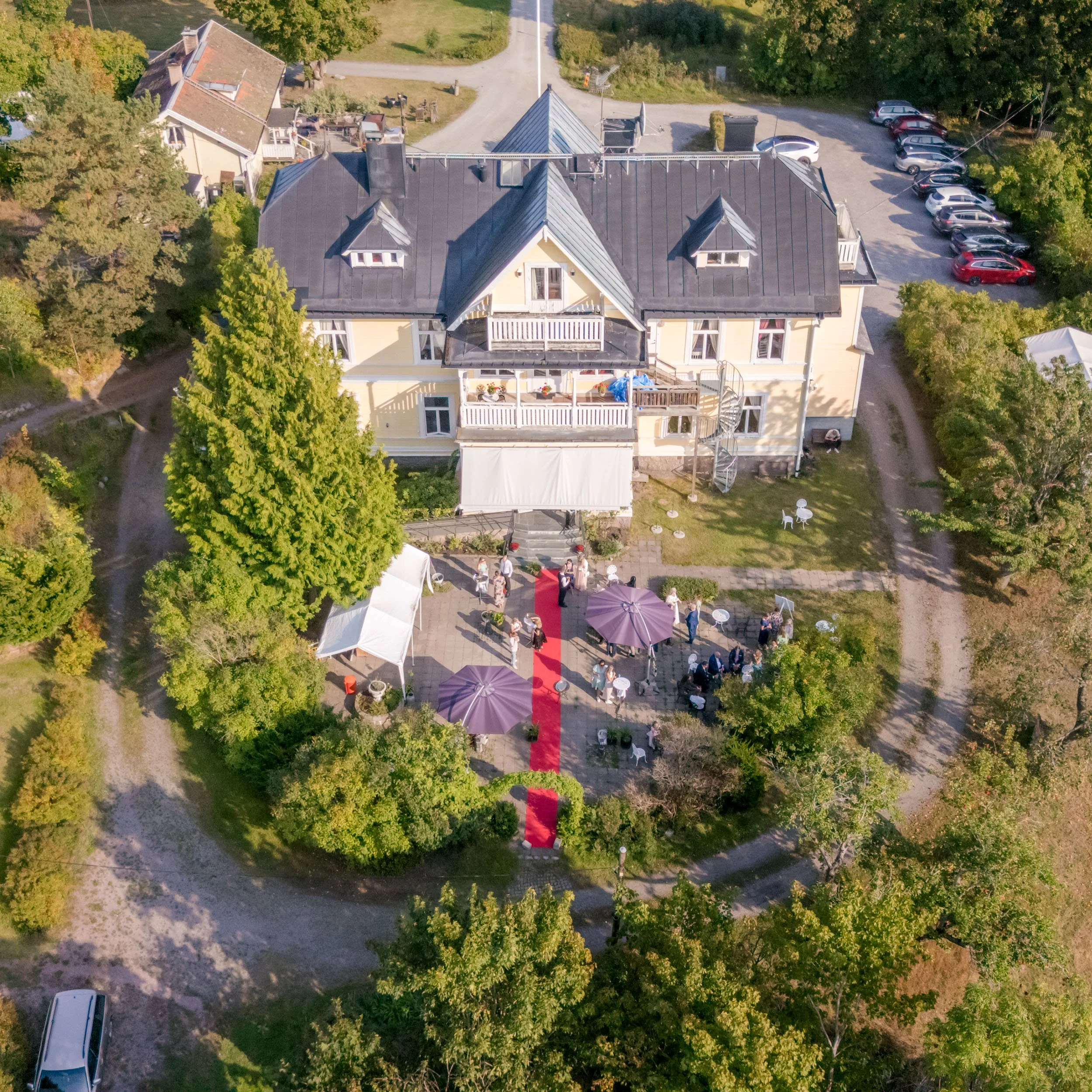 En stor ljusgul herrgård med mörk tak och många fönster, omgiven av träd och gröna områden. Utanför görs en del förberedelser för ett evenemang med gäster, bord, parasoller och röda mattor, samt ett tillfälligt tält.