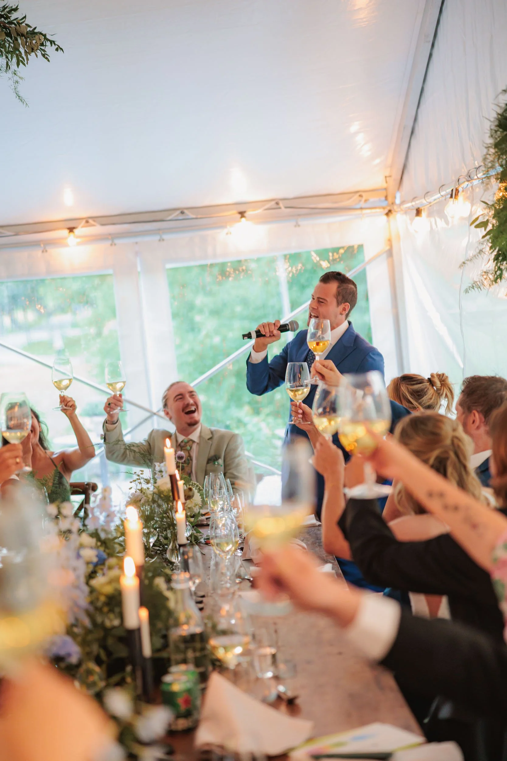Föreställer en festlig bröllopssammankomst med gäster som håller i glas med vin och skrattar. En man står med mikrofon och håller ett glas vin, medan andra runt bordet lyfter sina glas och skrattar.