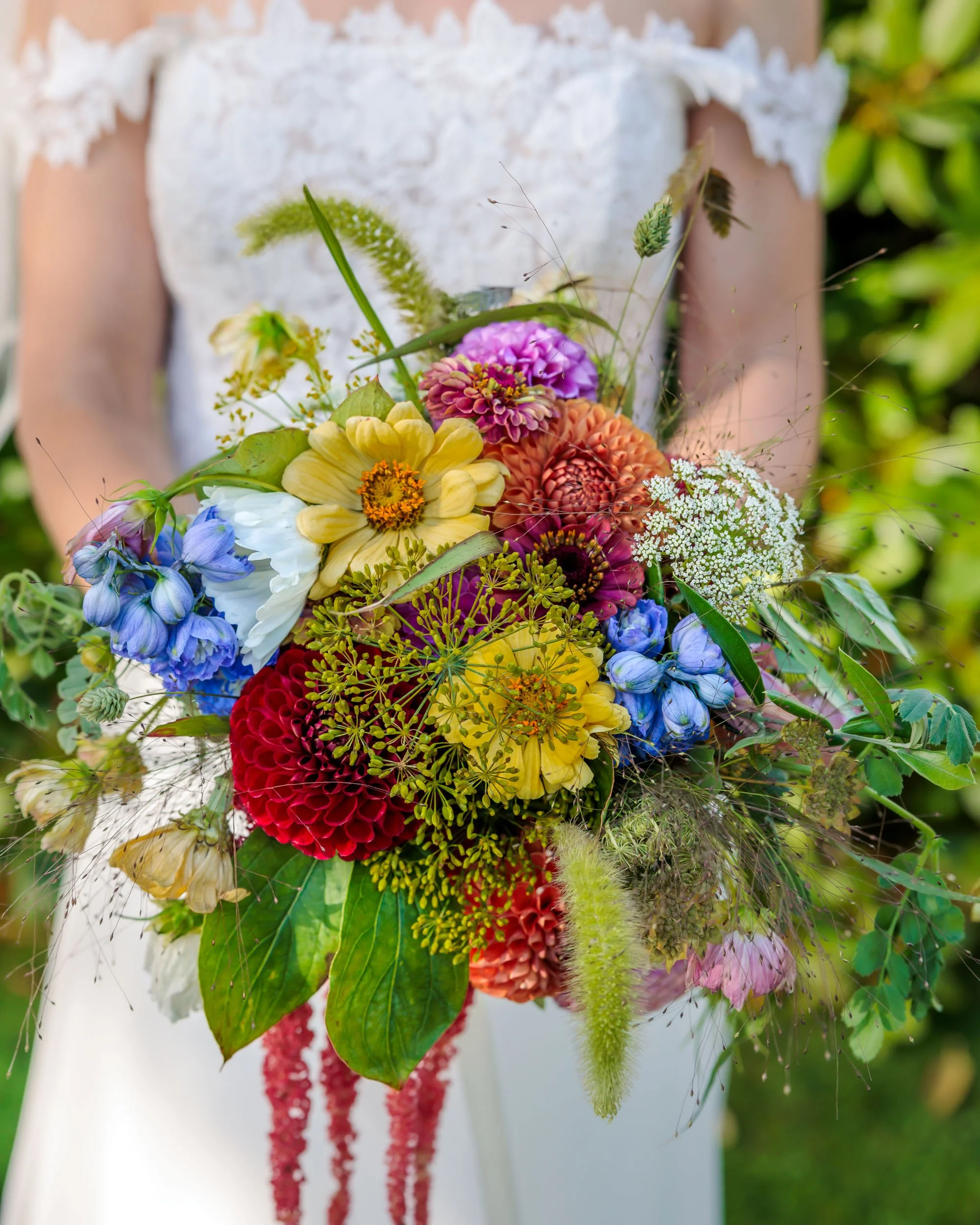 En brud i vitt plagg håller en färgglad brudbukett med olika blommor som ljusgult, rött, blått, rosa och vitt.