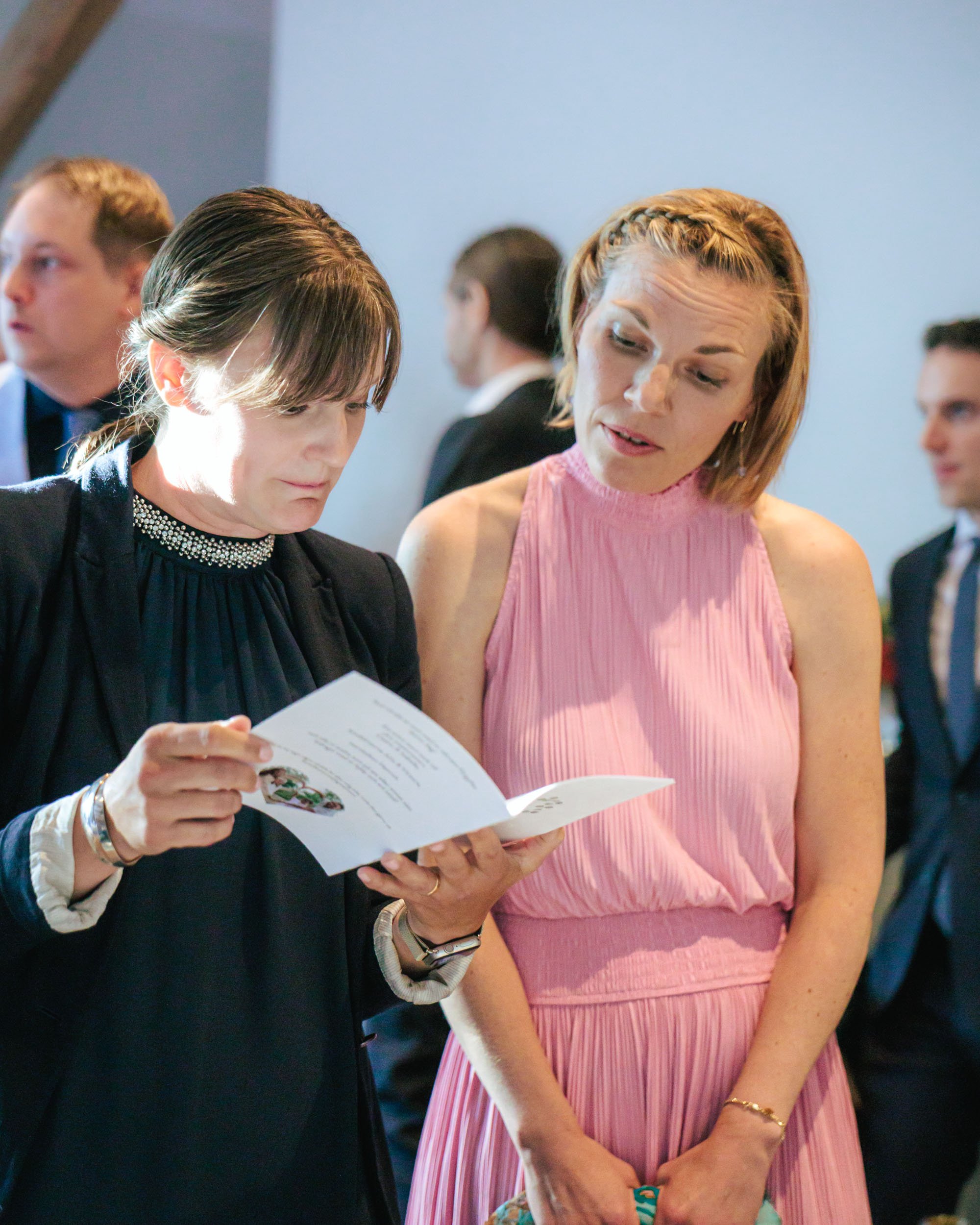 Två kvinnor i samtal, en håller ett programblad, på en formell tillställning med andra personer i bakgrunden.