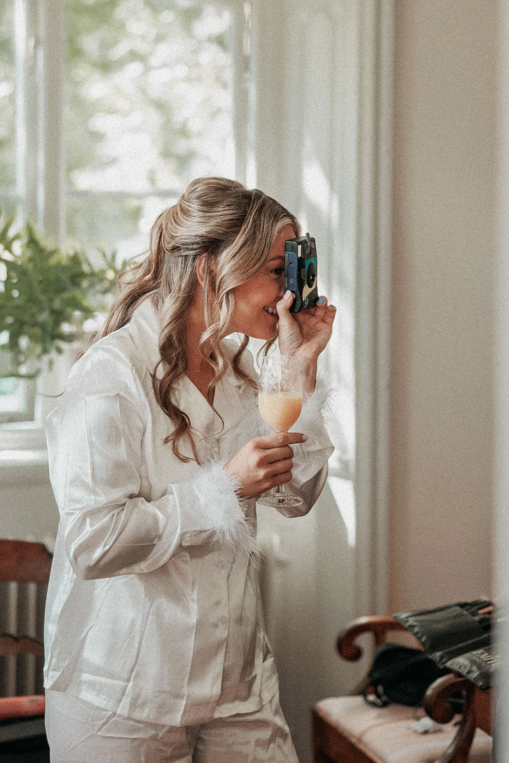 En kvinna i vit pyjamas fotograferar med en gammal kamera och håller ett glas champagne.