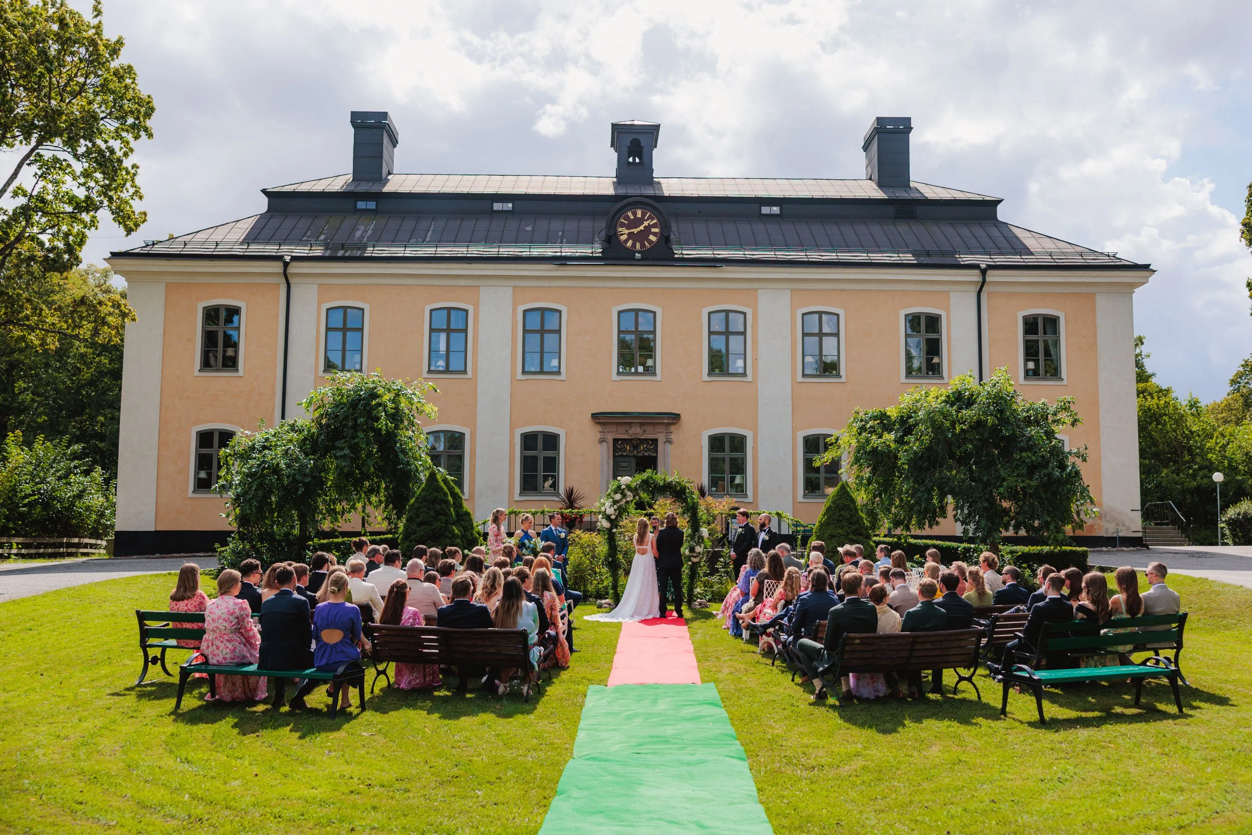 Bröllopsceremoni utomhus framför ett stort gult hus. Brud och brudgume står i centrum, omgivna av gäster sittande på bänkar. En röd matta leder till ceremoniplatsen, omgiven av gröna träd och buskar.