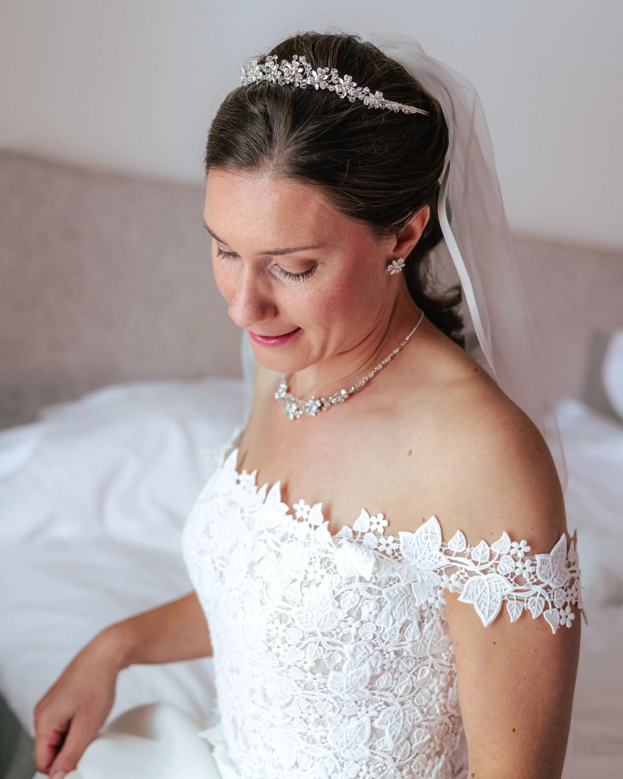 Brud i vit klänning med blommönster, smycken och en tiara, sittande på en säng.