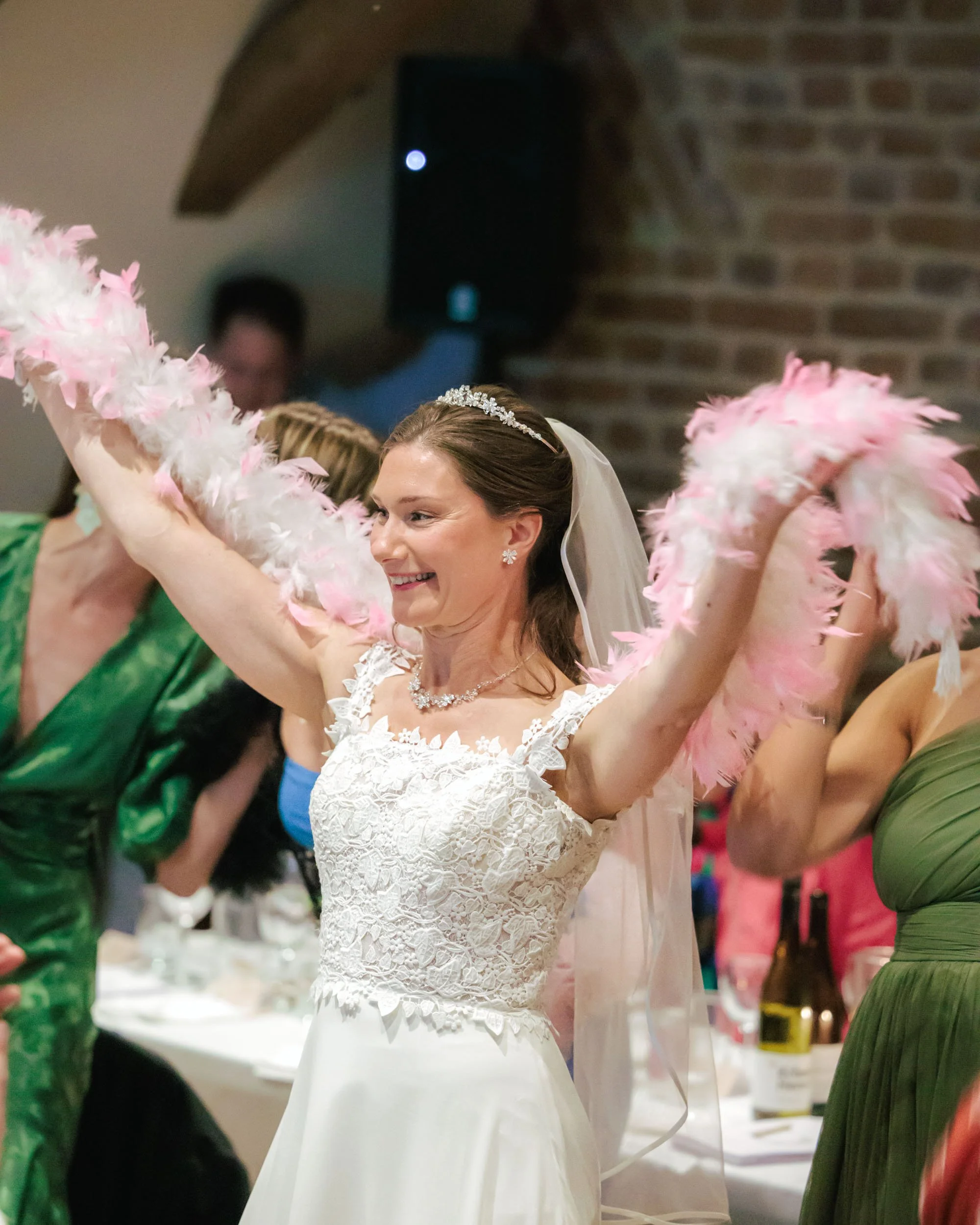 En brud i vit bröllopsklänning dansar med rosa och vita fjädrar på en fest, omgiven av gäster.