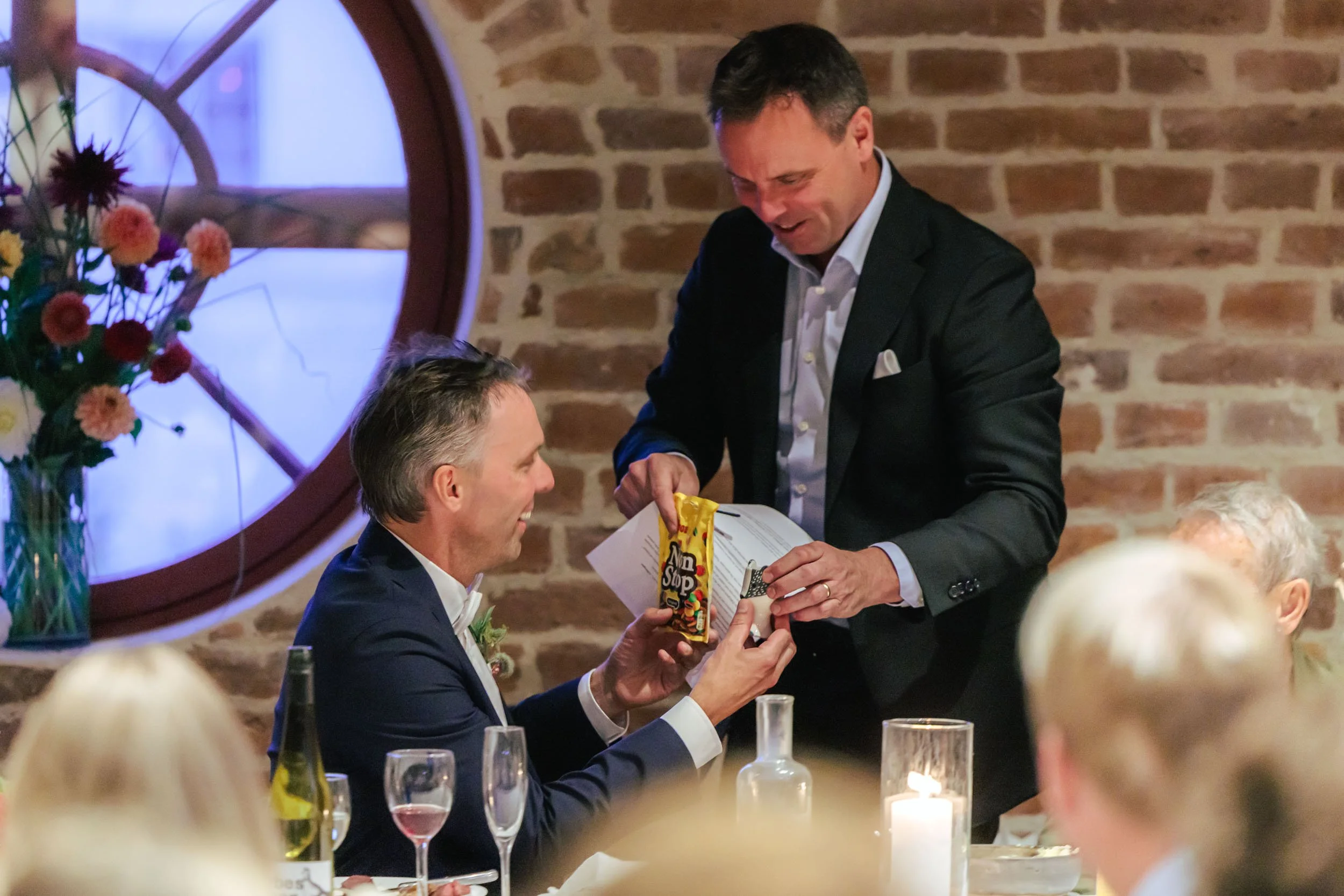 En man i mörk kostym som ger en present till en annan man i mörk kostym vid ett bord under en fest i ett rum med tegelväggar och ett runt fönster, omgiven av ljuslyktor och elegant dukning.