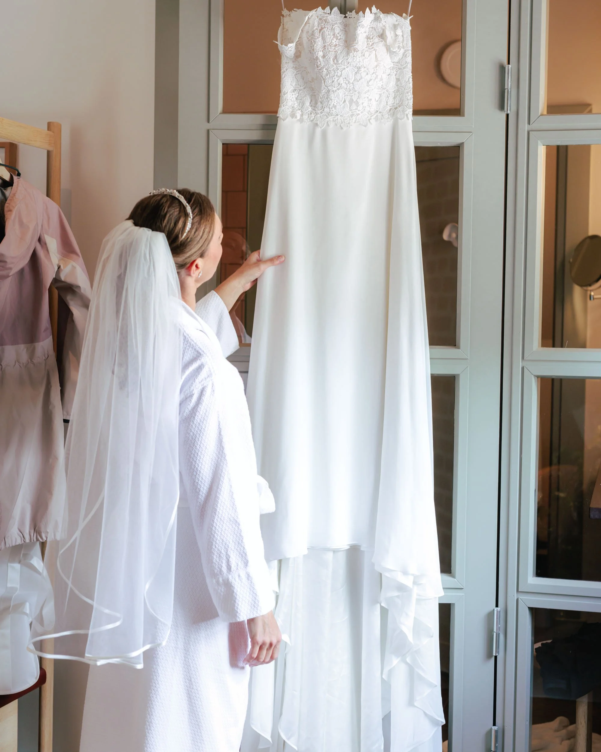 En brud i vit klänning med slöja som tittar på en vit brudklänning som hängs upp.
