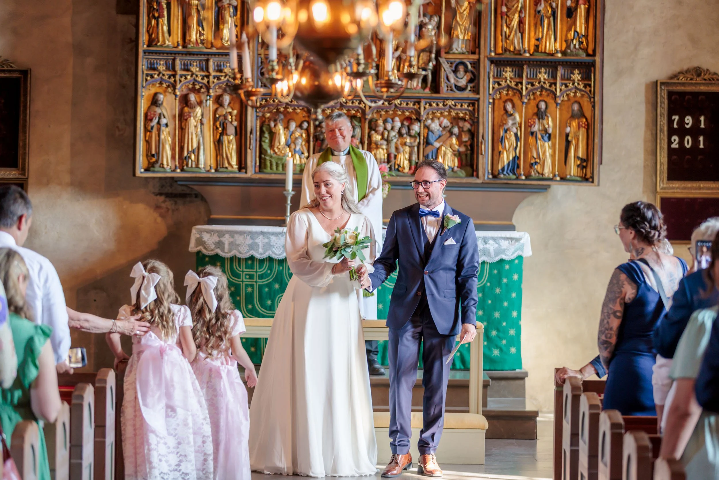 En brud och en brudgång skakar hand under ett bröllop i en kyrka, med en präst i bakgrunden och gäster på bänkar. Bruden bär vit klänning och håller en blomsterbukett, medan brudgummen har på sig en mörkblå kostym med fluga.