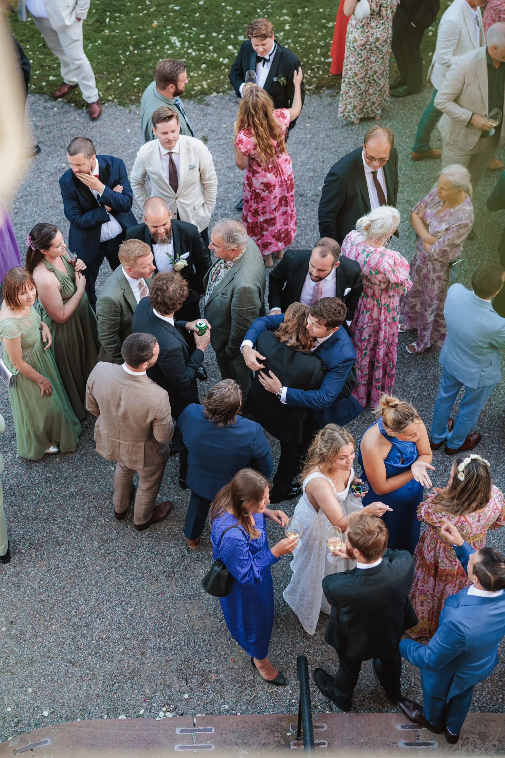 Folk samlade på ett utomhusbröllop, samtalar och firar, klädda i formella och färgglada kläder.