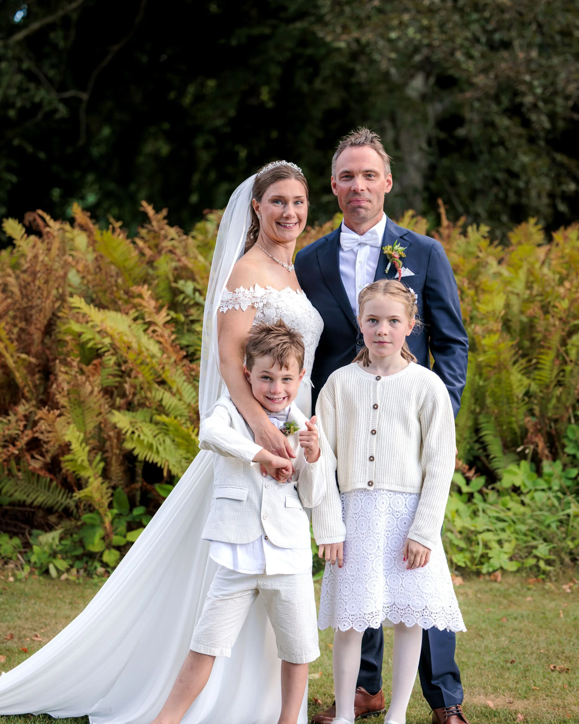 Ett brudpar med två barn utomhus, med gröna buskar och träd i bakgrunden, vid ett bröllop.