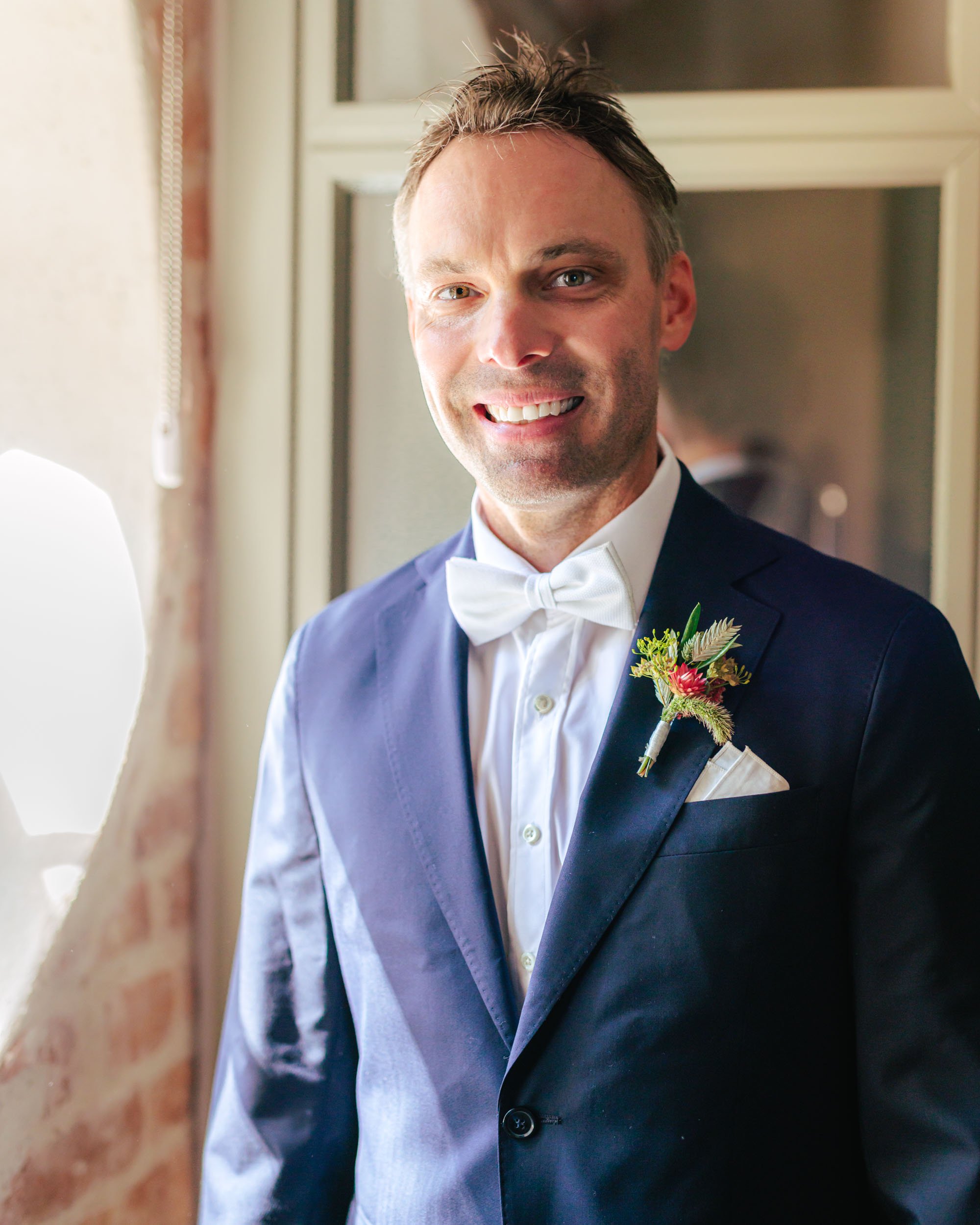 En glad man i bröllopskläder, vit fluga, vit näsduk och blommor i kavajslaget, står vid ett fönster inomhus.