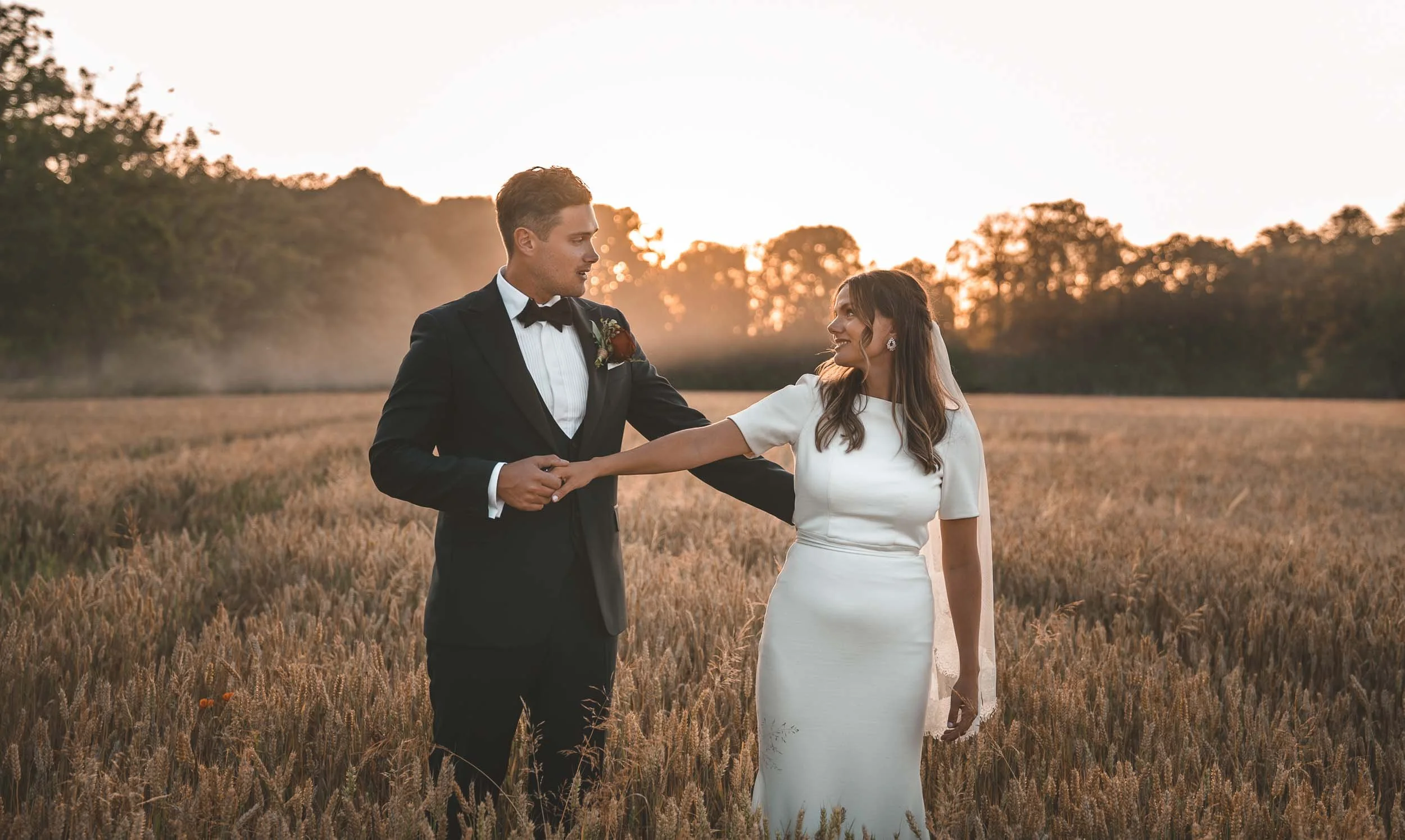En man och en kvinna i bröllopskläder står i ett fält av vete under en solnedgång, håller varandras händer och tittar på varandra.