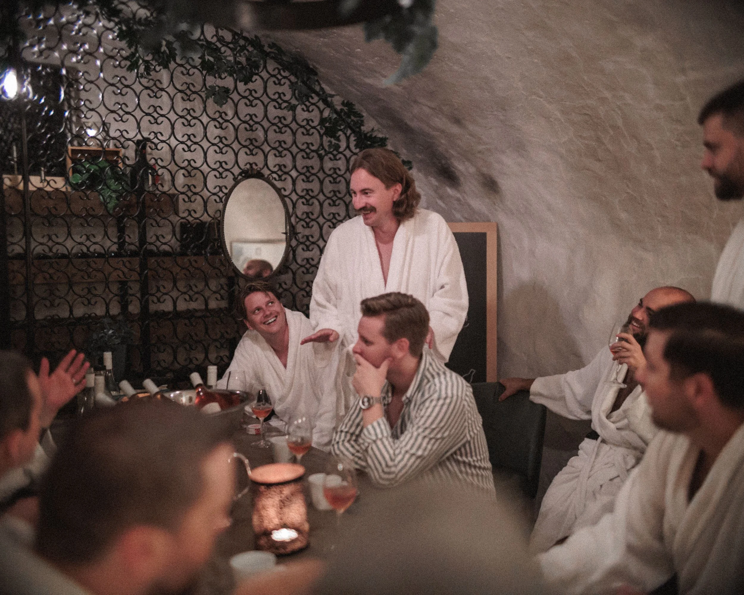 Flera personer i badrock samlas runt ett bord i ett mysigt rum med bärnstensfärgad belysning, och en man med långt hår och skägg talar och ler.