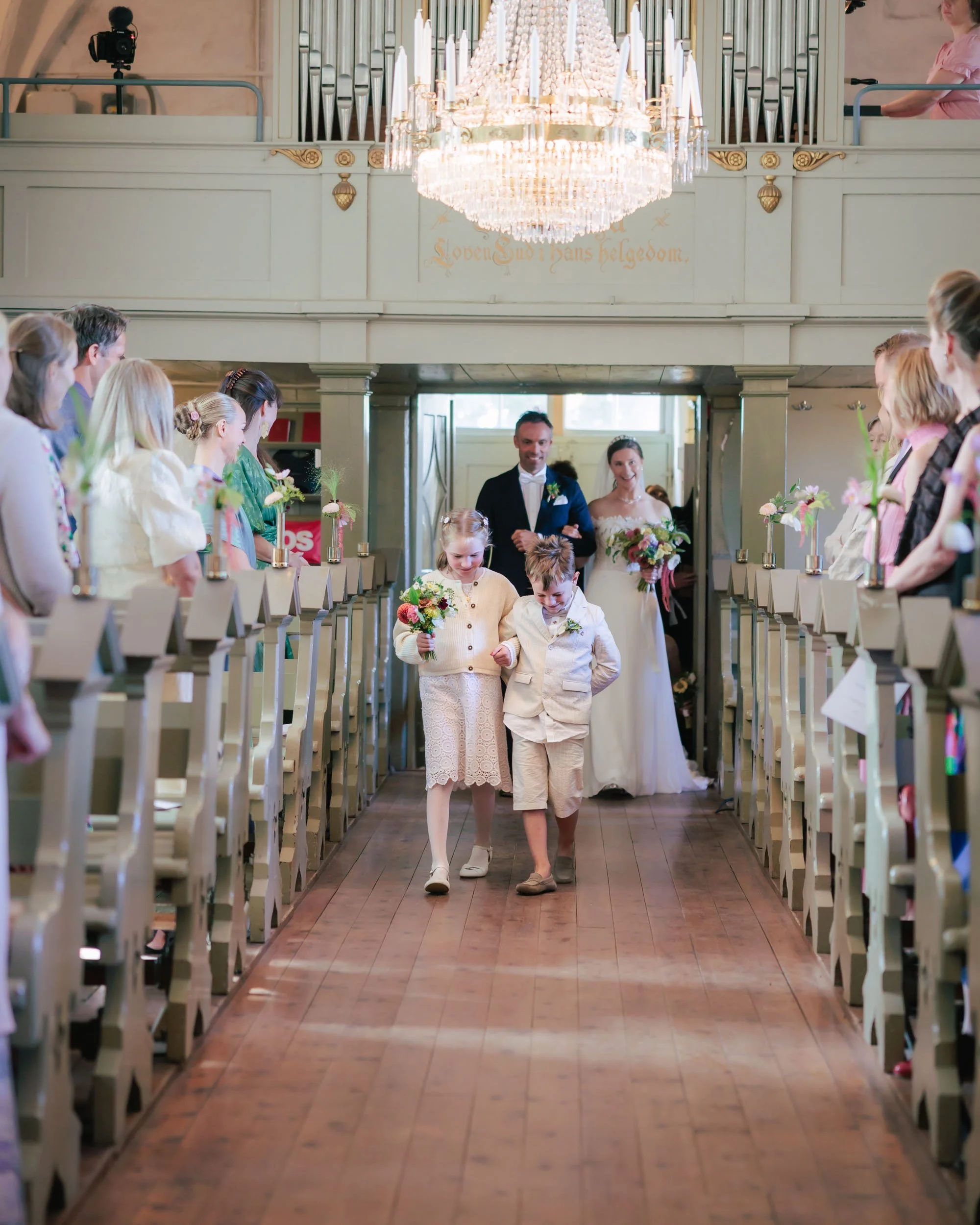 Par i bröllopskläder går nerför kyrkans gång, följda av ett barnpar, medan gäster ser på.