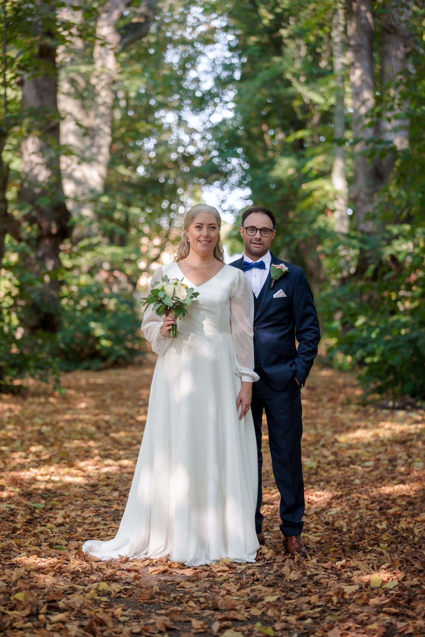 Brudpar står i en skog på bröllopsdag, kvinnan bär en vit brudklänning och håller en bukett, mannen bär en mörk blå kostym och har en blå fluga.