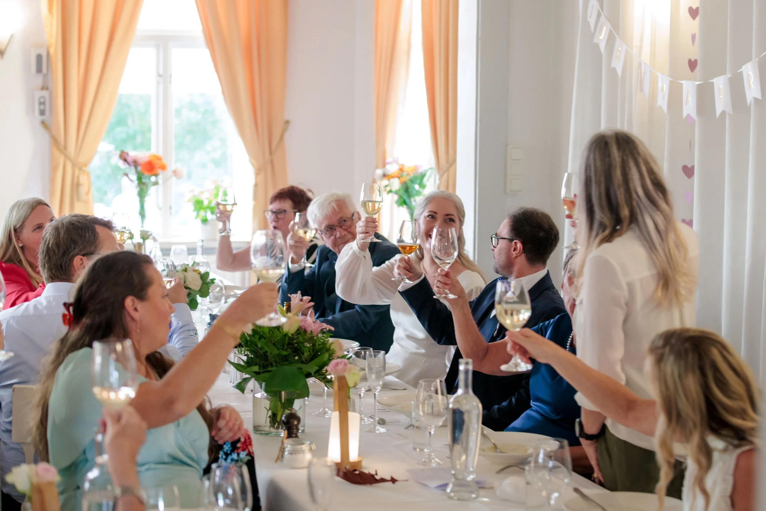 Människor som samlas runt ett bord och skålar med glas vin vid en festlig tillställning i ett vardagsrum med fönster och gardiner.