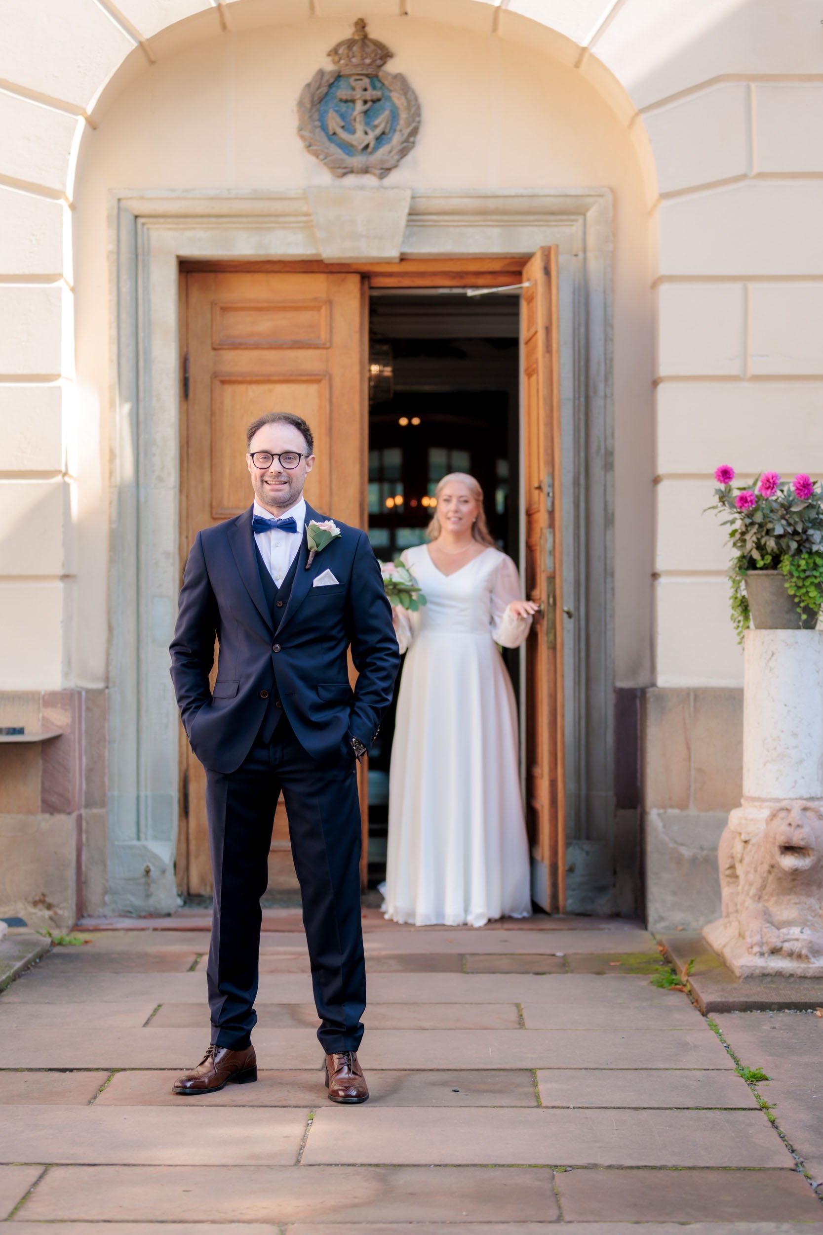 En man i mörk kostym och röd slips står framför ingången till en byggnad, med en kvinna i vit brudklänning som tittar ut genom dörren bakom honom.