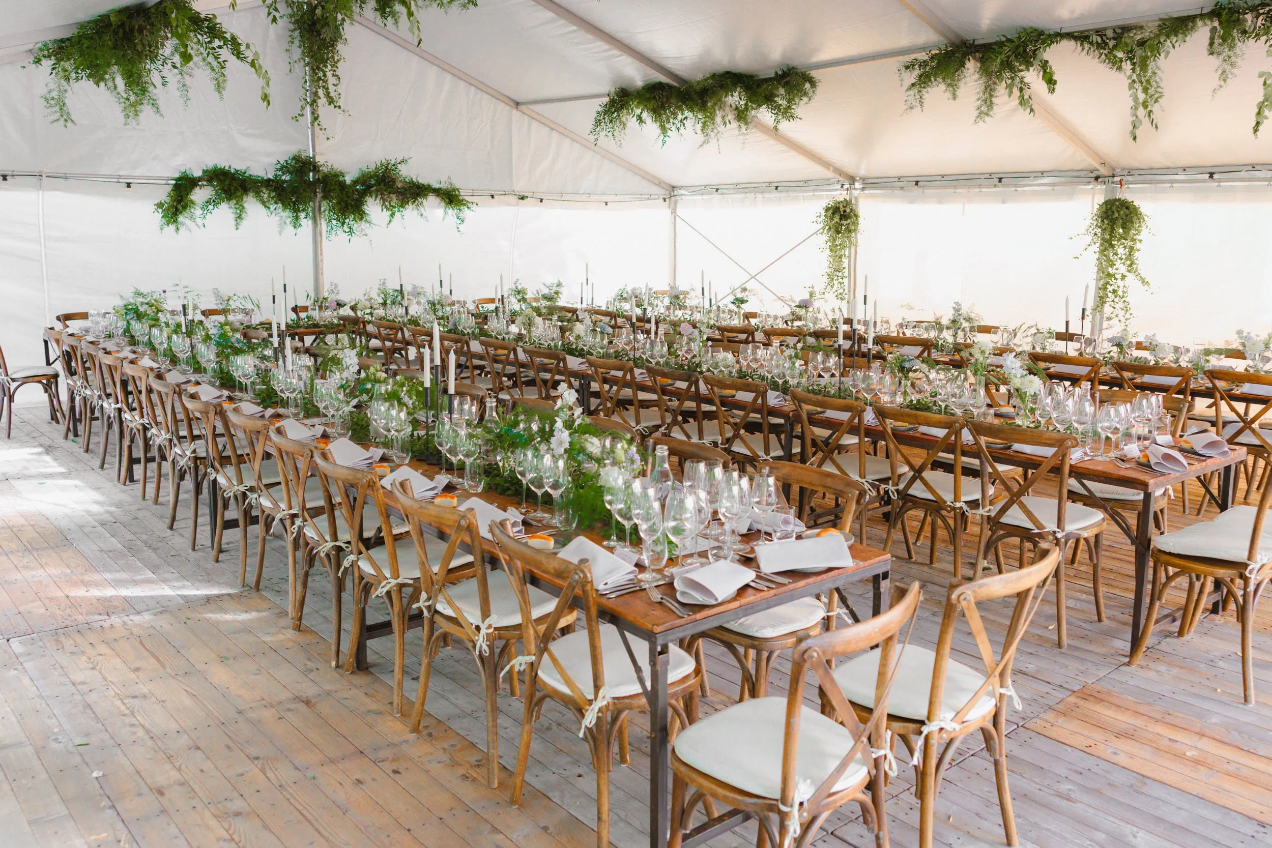 Festmiddagsbord i ett tält med träbord, vita dukar, glas, tallrikar, servetter och gröna växter, med gröna växter som dekorationer.