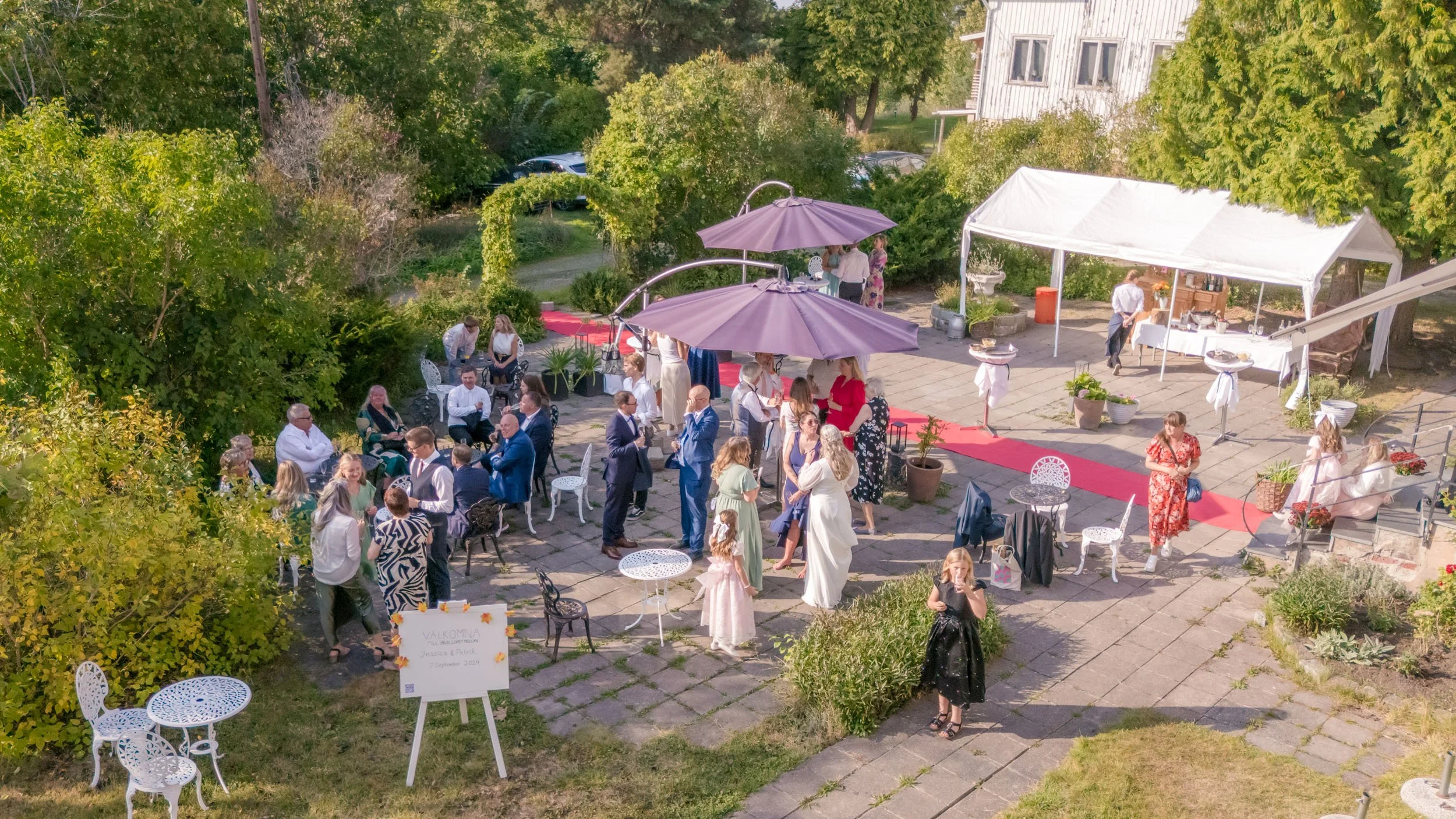 Förrättsskylt med välkomnste till bröllopsfesten, många gäster minglar, sitter och står på en utomhusfest under parasoller och tält, med ett rött mattband på golvet.