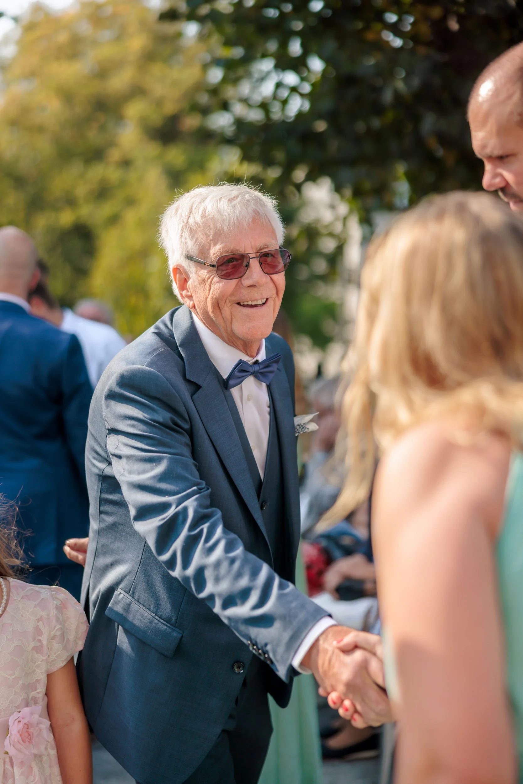 En äldre man i kostym som ler och skakar hand med en kvinna på ett utomhusevent, andra människor syns i bakgrunden.