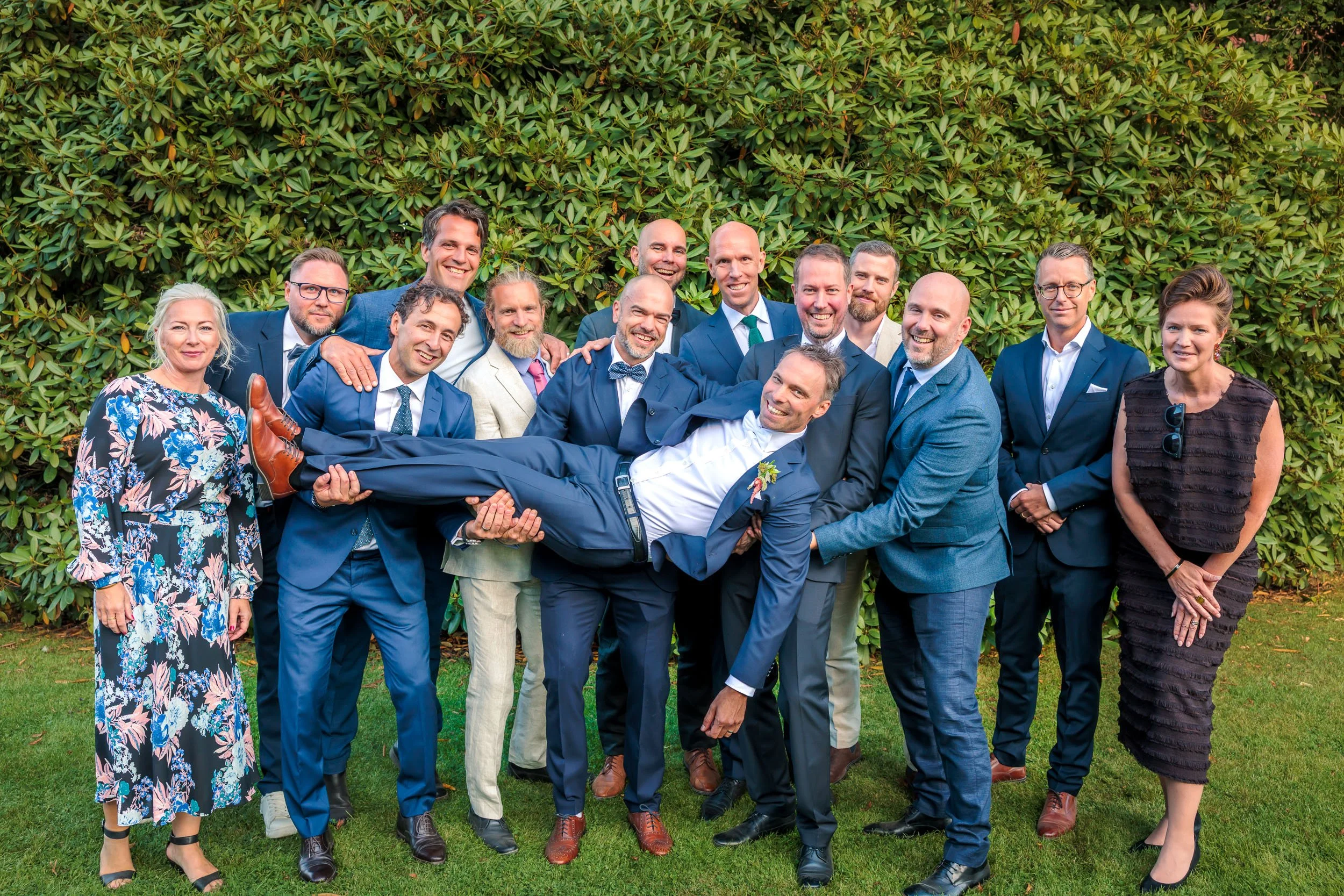 Grupp av personer i formella kläder, en man hålls horisontal av andra, utomhus framför ett grönt buskage, firar ett bröllop.