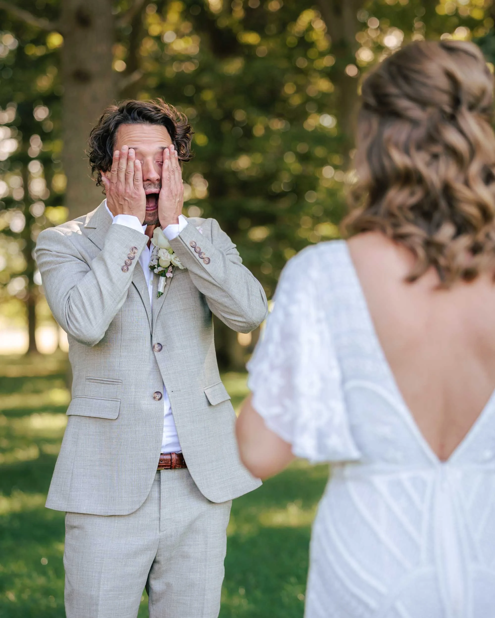 A man in a light-colored suit is crying tears of joy and surprise at a wedding, looking at a woman in white standing in front of him in a lush park.