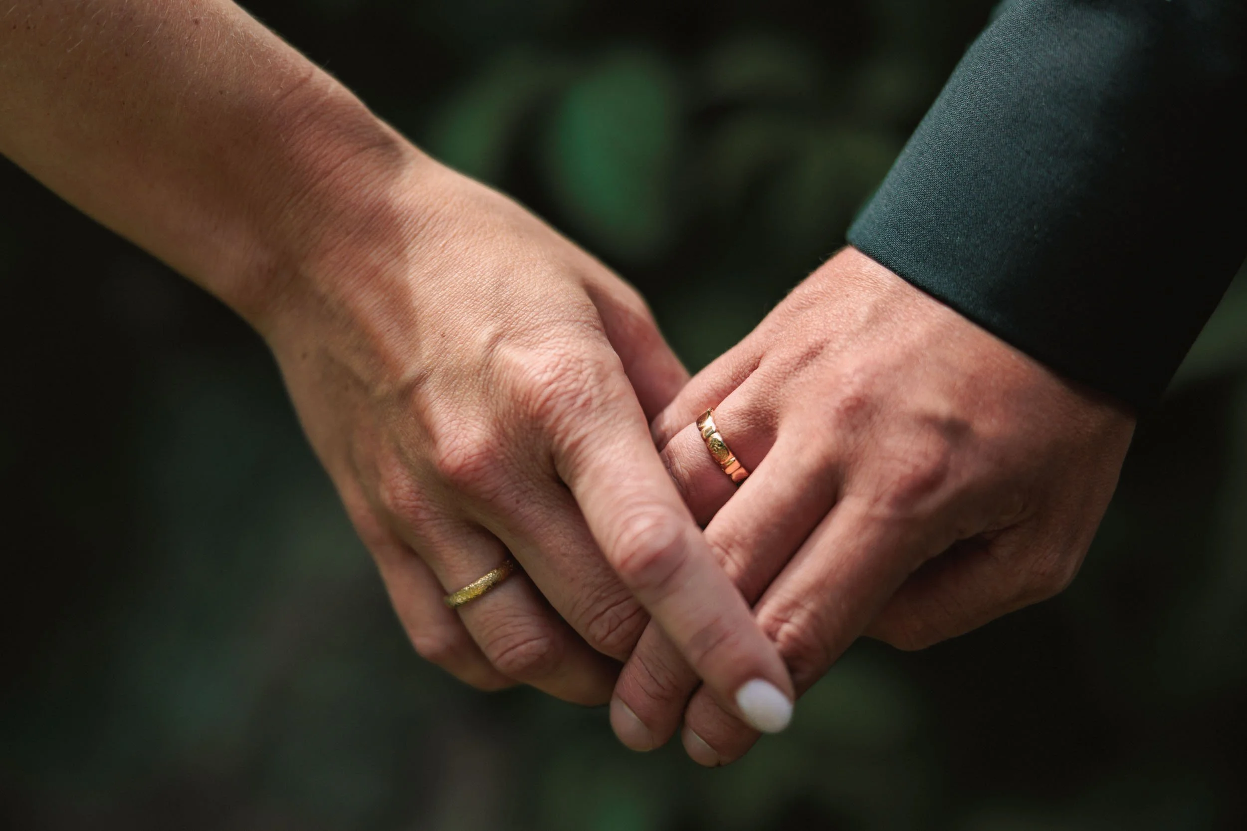 Två personer håller om varandra med ringar på fingrarna, en person i mörk dräkt och den andra i ljus hudton. Bakgrund är suddig och mörk.