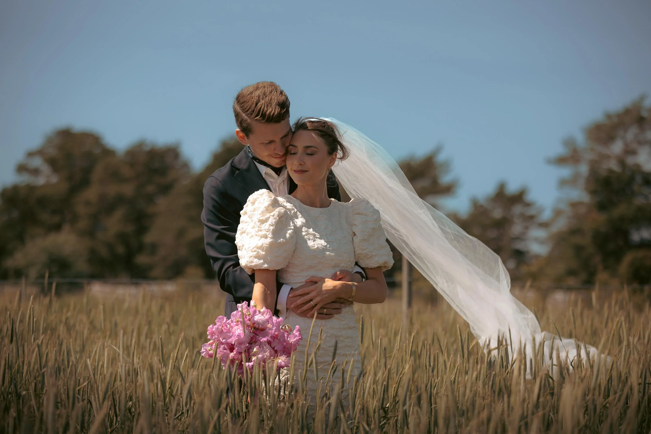 En brud och en brudgum står i ett fält av höstgräs, bruden bär en vit klänning med puffiga ärmar och ett släp av tyll, med en lång slöja över huvudet. Brudgummen iklädd en svart kostym håller om henne. Hon håller en bukett med rosa blommor.
