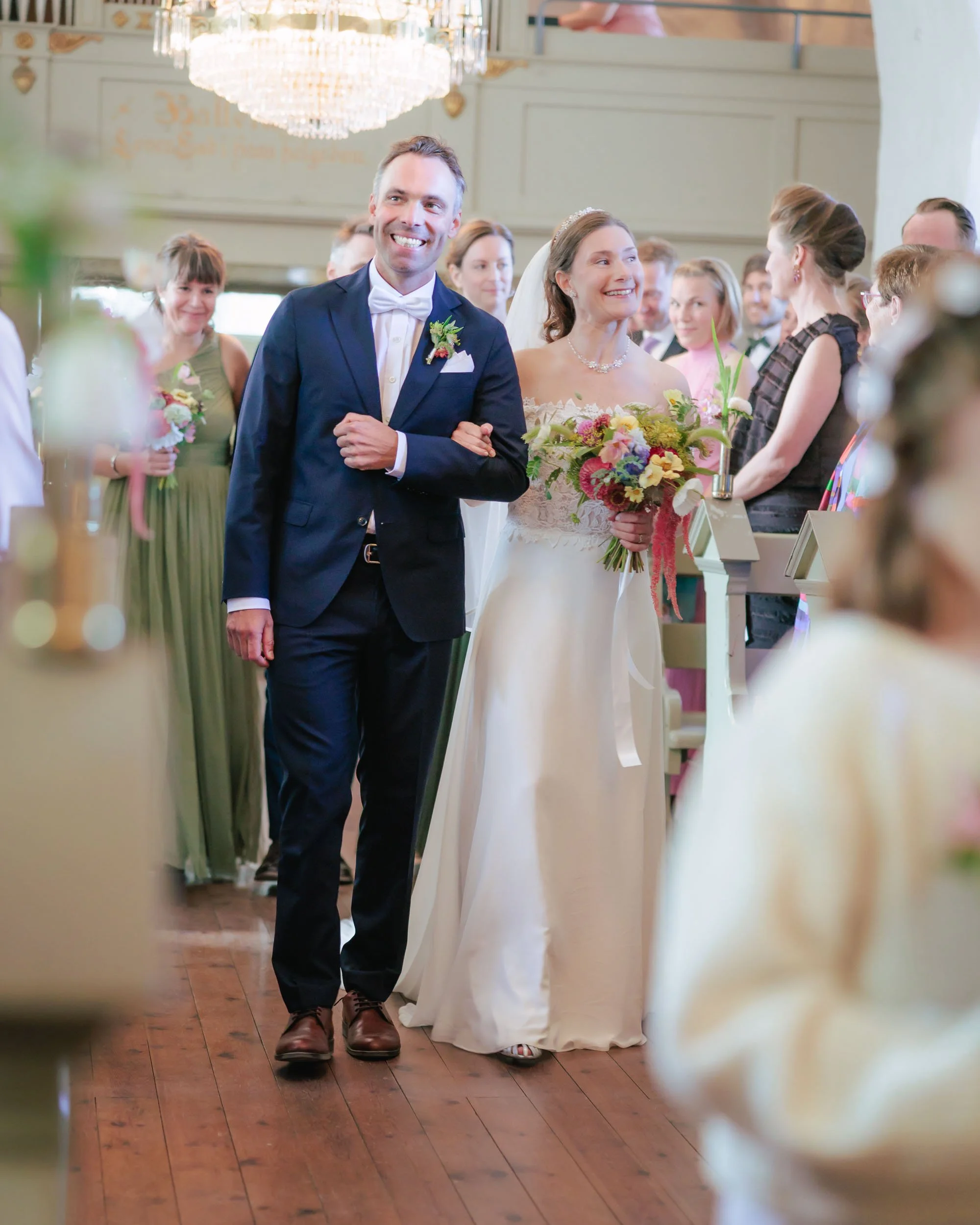 Brudparet med mannen i mörk kostym och kvinnan i vit brudklänning, som är på väg in i ett bröllopsceremoni. Kvinnan håller en färgglad bukett och båda ler.