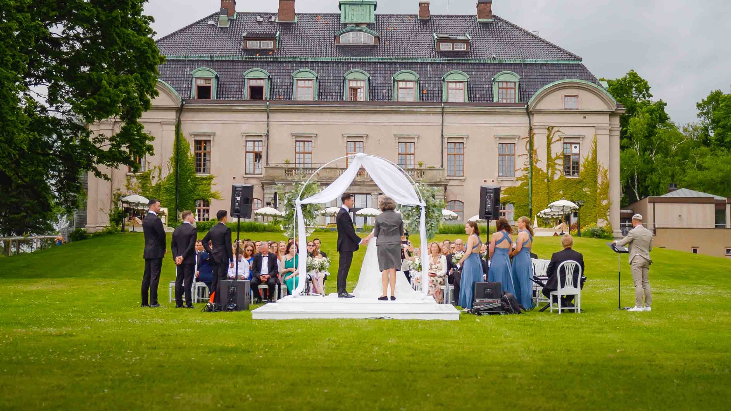 Bröllopsceremoni utomhus framför ett stort slott, med gäster som sitter i rad och brudparet under ett vitt draperat valv.