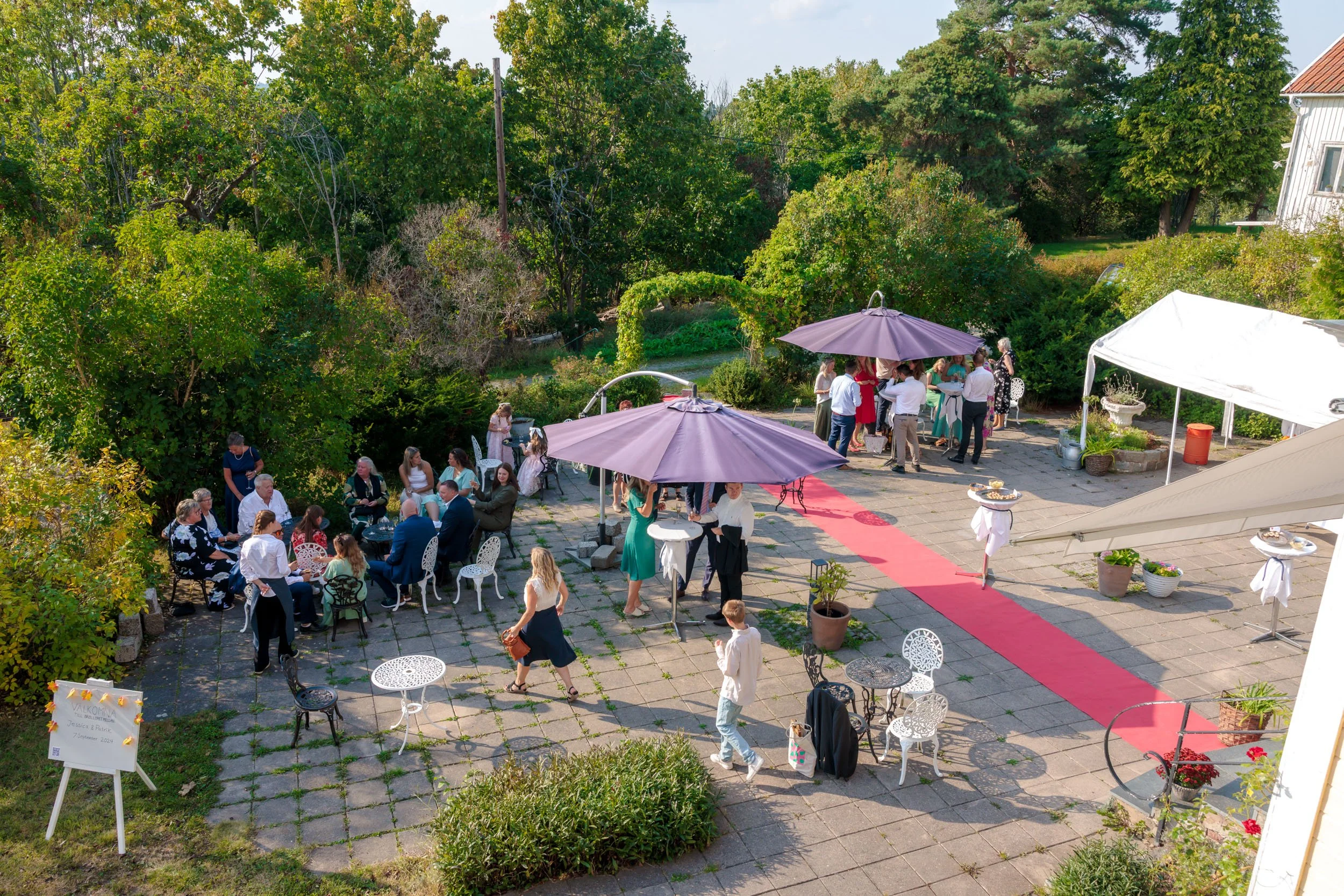 Utsikt över ett utomhus party med flera små grupper av människor, som minglar och äter under parasoller, på en stenbelagd trädgårdsplaner, omgiven av gröna träd och buskar, med en röd matta på marken.